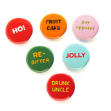 An inverted triangle of six holiday christmas pinback buttons. Left to Right starting on the top row: Ho! a red button with white text, and an exclamation point at the end of Ho! Fruit Cake a golden brown button with FRUIT CAKE written in red and green candy swirls. GAY APPAREL is a pink button with light green text. RE-GIFTER is a green button with yellow-gold text.  JOLLY is a mint green button with red text.  And lastly at the bottom is DRUNK UNCLE a red button with lime green text.