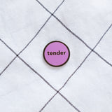 Round enamel pin that says TENDER on fabric background.