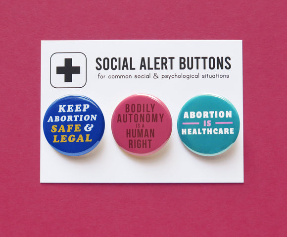 Three round pinback buttons lined up on a SOCIAL ALRT BUTTONS backing card. Left to right: Blue button reads Keep Abortion Safe & Legal, Middle pink-red button reads Bodily Autonomy is a Human Right, and last teal button reads Abortion is Healthcare.