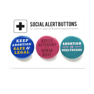 Three round pinback buttons lined up on a SOCIAL ALRT BUTTONS backing card. Left to right: Blue button reads Keep Abortion Safe & Legal, Middle pink-red button reads Bodily Autonomy is a Human Right, and last teal button reads Abortion is Healthcare.