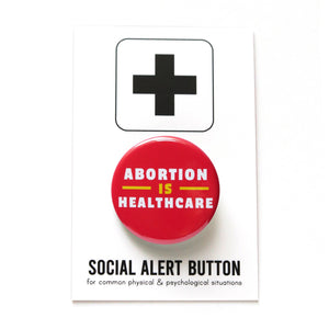 A round teal-blue pinback button that reads ABORTION IS HEALTHCARE in three lines in white & lavender text. Badge is pinned to a Social Alert Button backing card, with a plus sign at the top.