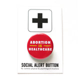 A round red pinback button that reads ABORTION IS HEALTHCARE in three lines in white & yellow text. Badge is pinned to a Social Alert Button backing card, with a plus sign at the top.