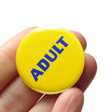 Round pinback button that says ADULT. Blue text on a yellow background. The button is held in a hand.