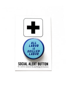 Round light blue pinback button with a dark blue outline & dark blue text that reads ALL LABOR IS SKILLED LABOR. The button is on a rich warm caramel colored sweatshirt.
