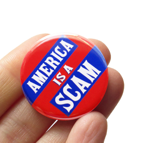 A round red pinback button with blue rectangles to pop white text that reads AMERICA IS A SCAM. Button is held in a hand.