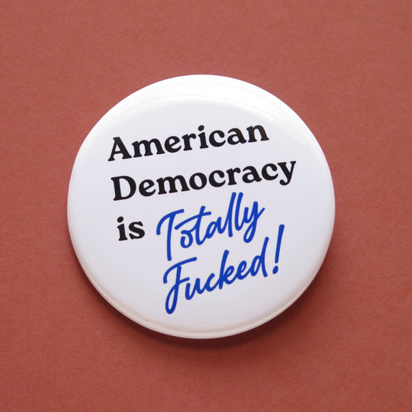 A round white pinback button that reads American Democracy is Totally Fucked in black and blue text. Button is on a clay-red background.