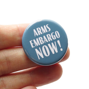 A round gray blue pinback button that reads ARMS EMBARGO NOW! in white text. Button is held in a hand.