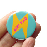 Round 1.25" aqua pinback button that reads ART FAG in pink text on a yellow lightening bolt. Badge is held in hand.