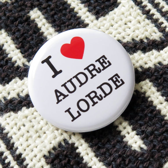 Round white pinback button that reads I Love Audre Lorde, with love indicated with a red heart. Pinned to a black and white woven jacket.