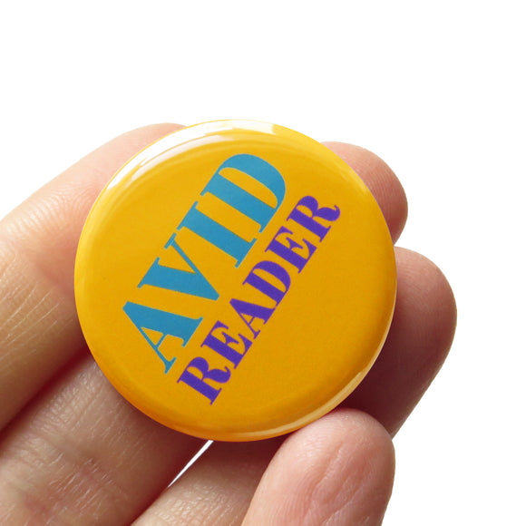 Round yellow colored pinback button which reads Avid Reader in pruple and teal text. The button is held in a hand.