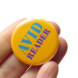 Round yellow colored pinback button which reads Avid Reader in pruple and teal text. The button is held in a hand.