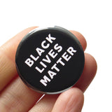 Round pinback button that says BLACK LIVES MATTER. White, text on a black background. Button is held in a hand.