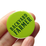 Neon green round pinback button, which reads BACKYARD FARMER in a dark green font. Button is held in a hand