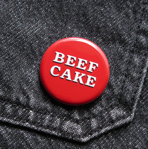 A red pinback button that reads BEEF CAKE in two lines in white serif text with a black drop shadow. Button is pinned to black denim