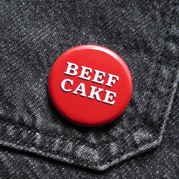 A red pinback button that reads BEEF CAKE in two lines in white serif text with a black drop shadow. Button is pinned to black denim