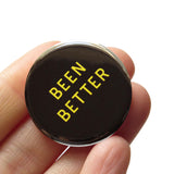 A round black pinback buton that reads BEEN BETTER in yellow text. Badge is held in a hand.