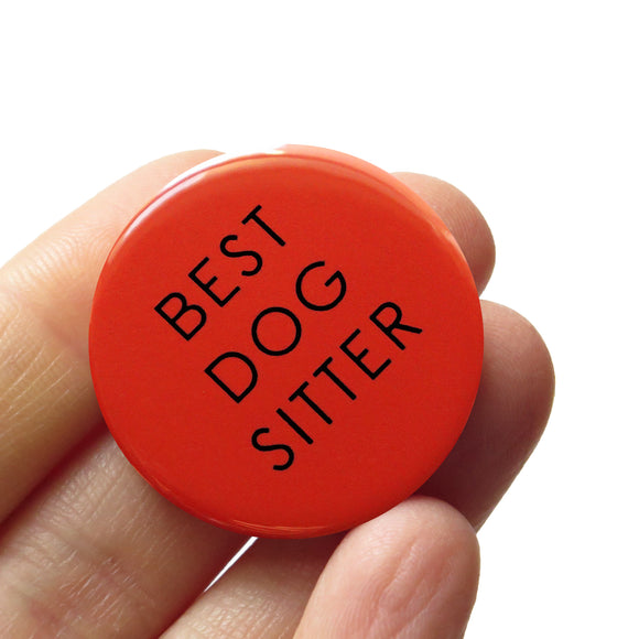 Round orange-brown pinback button that reads BEST DOG SITTER in a thin black font. Button is held in a hand.