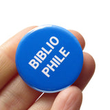 Round bright blue colored pinback button which reads BIBLIOPHILE in a white sans serif font on two lines. The button is held in a hand.