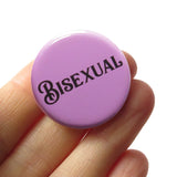 Round lavender pinback button that reads BISEXUAL in black script font. Held in a white hand.