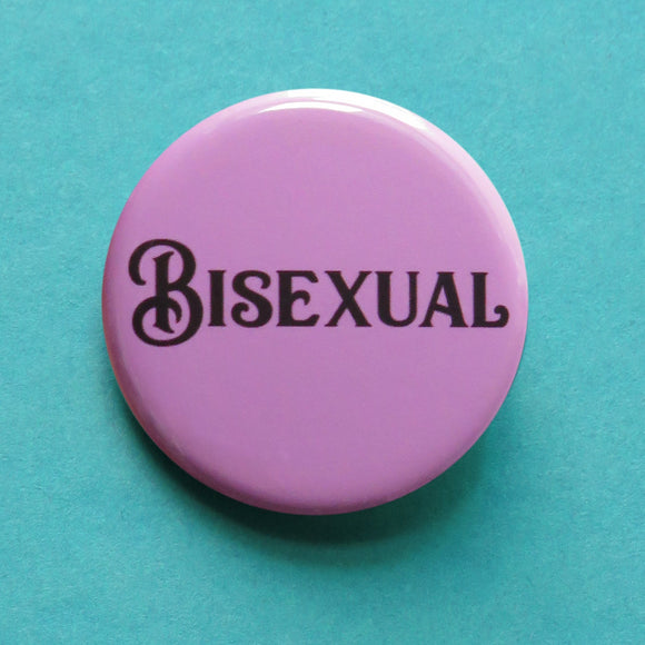 Round lavender pinback button that reads BISEXUAL in black script font. The button is on a teal background