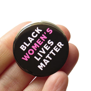 Round black pinback button that reads BLACK WOMEN'S LIVES MATTER, in white with Women's in pink for emphasis. Button is held in a hand.