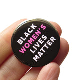 Round black pinback button that reads BLACK WOMEN'S LIVES MATTER, in white with Women's in pink for emphasis. Button is held in a hand.