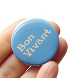 Round, light blue pinback button with cream text that reads BON VIVANT on two lines. Badge held in a hand.