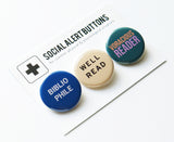 Three round pinback buttons lined up on a SOCIAL ALERT BUTTONS backing card, angled. Reading left to right: A round blue button that reads BIBLIOPHILE in white text on two lines, A cream button that reads WELL READ in black text on two lines, and a teal button that reads VORACIOUS READER in peach & lavender on two lines.