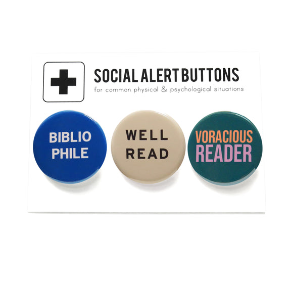 Three round pinback buttons lined up on a SOCIAL ALERT BUTTONS backing card. Reading left to right: A round blue button that reads BIBLIOPHILE in white text on two lines, A cream button that reads WELL READ in black text on two lines, and a teal button that reads VORACIOUS READER in peach & lavender on two lines.