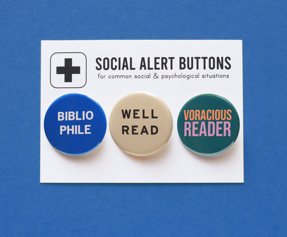 Three round pinback buttons lined up on a SOCIAL ALERT BUTTONS backing card. Reading left to right: A round blue button that reads BIBLIOPHILE in white text on two lines, A cream button that reads WELL READ in black text on two lines, and a teal button that reads VORACIOUS READER in peach & lavender on two lines.