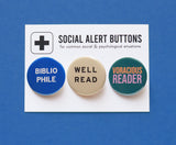 Three round pinback buttons lined up on a SOCIAL ALERT BUTTONS backing card. Reading left to right: A round blue button that reads BIBLIOPHILE in white text on two lines, A cream button that reads WELL READ in black text on two lines, and a teal button that reads VORACIOUS READER in peach & lavender on two lines.