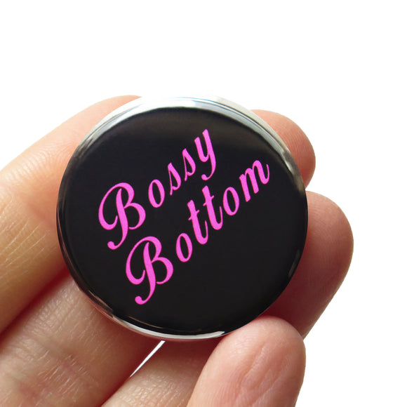 Round pinback button that reads BOSSY BOTTOM in bright pink text on a black background. Button is held in a hand.