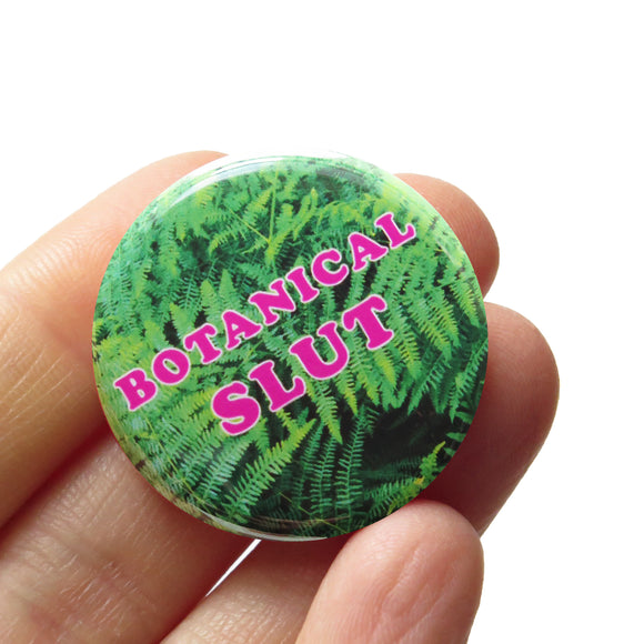 Round pinback button that says BOTANICAL SLUT. Pink text on a fern photo background. Badge is held in a hand.