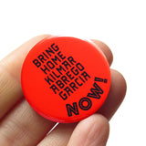 A round red-orange 1.25" pinback button that reads BRING HOME KILMAR ABREGO GARCIA NOW! in black text. Button is held in a hand.