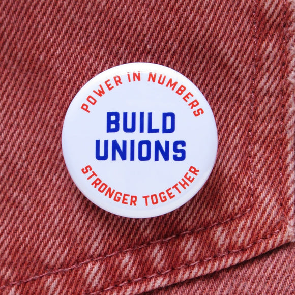 Around white button that reads BUILD UNIONS in bold text in the center. The upper curve reads: Power in Numbers, in orange text, The lower curve reads, Stronger Together in orange text. Button is pinned to an orange denim pocket.