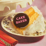 Round chocolate brown pinback button that reads Cake Baker in medium pink san serif font. Button is on top of a vintage cookbook photo of a piece of cake.