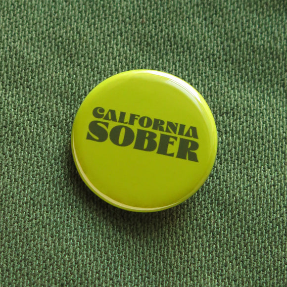 Round yellow green pinback button that says CALIFORNIA SOBER in dark green text. Button is pinned to a dark green cotton fabric.