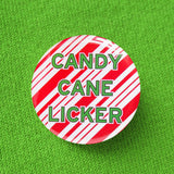 Round pinback button that says CANDY CANE LICKER in light green text on a red candy cane striped background. Button is pinned to a moss lime green sweatshirt