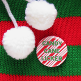 Round pinback button that says CANDY CANE LICKER in light green text on a red candy cane striped background. Button is on a Social Alert Button backing card. Button is pinned toa red and white knit stocking with two puffy white pom pom balls.