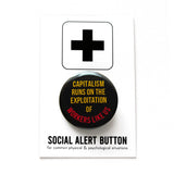 A round black 1.25" pinback button that reads Capitalism sRuns on the exploitation of workers like us in red and yellow text. Badge is pinned to a Social Alert Button backing card.