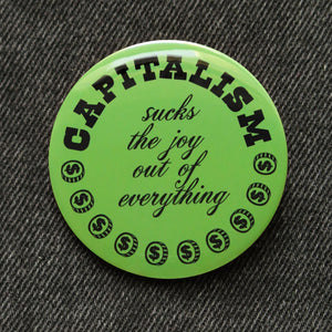 A round moss green pinback button of medium size that reads CAPITALISM in chunky black test at the top and in a black script in the middle: Sucks The Joy Out of Everything. Coins with a dollar sign fill a half moon around the edge of the button. Button is on black denim