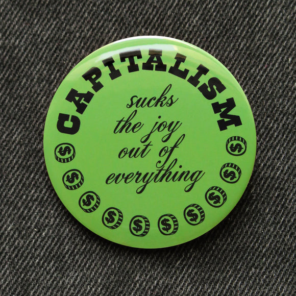 A round moss green pinback button of medium size that reads CAPITALISM in chunky black test at the top and in a black script in the middle: Sucks The Joy Out of Everything. Coins with a dollar sign fill a half moon around the edge of the button. Button is on black denim