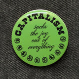 A round moss green pinback button of medium size that reads CAPITALISM in chunky black test at the top and in a black script in the middle: Sucks The Joy Out of Everything. Coins with a dollar sign fill a half moon around the edge of the button. Button is on black denim