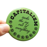 A round moss green pinback button of medium size that reads CAPITALISM in chunky black test at the top and in a black script in the middle: Sucks The Joy Out of Everything. Coins with a dollar sign fill a half moon around the edge of the button. Button is held in a hand.