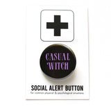 Round black pinback button that reads CASUAL WITCH in a lavender vintage 70’s font. The button is on a SOCIAL ALERT BUTTON backing card.
