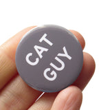 Round medium gray 1.25" pinback button that reads CAT GUY in white text . Badge is held in hand.