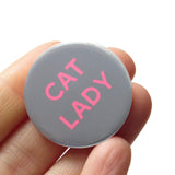 A round gray pinback button hat reads CAT LADY in pink text. Badge is held in a hand.