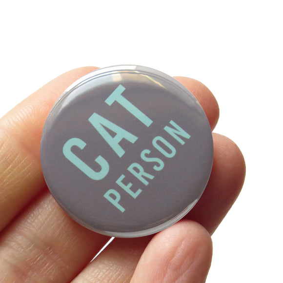 Round gray pinback button that reads CAT PERSON in mint green san serif text. Button is held in a hand.