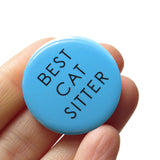 Round bright medium blue pinback button that reads BEST CAT SITTER in a thin black san serif text. Button is in a hand.