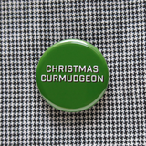 Round, dark green pinback button that says CHRISTMAS CURMUDGEON in white text with a black shadow. Button is on a tiny herringbone pattern shirt.